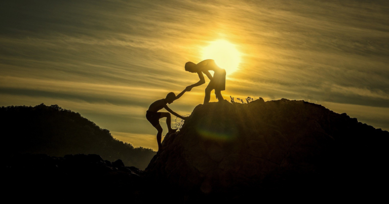 Two boys climbing up a large hill. The boy at the top has extended his hand to help the other boy up.