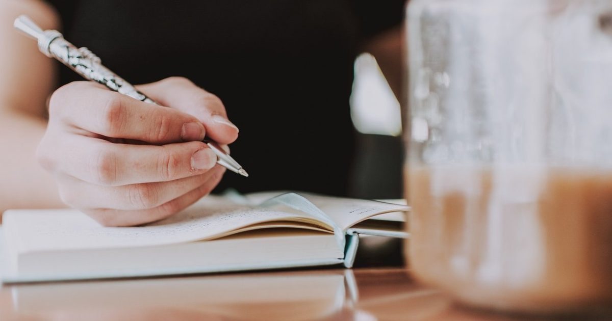 VIew of a woman's hand holding a pen with an open journal, a jar of iced coffee is in the foreground.