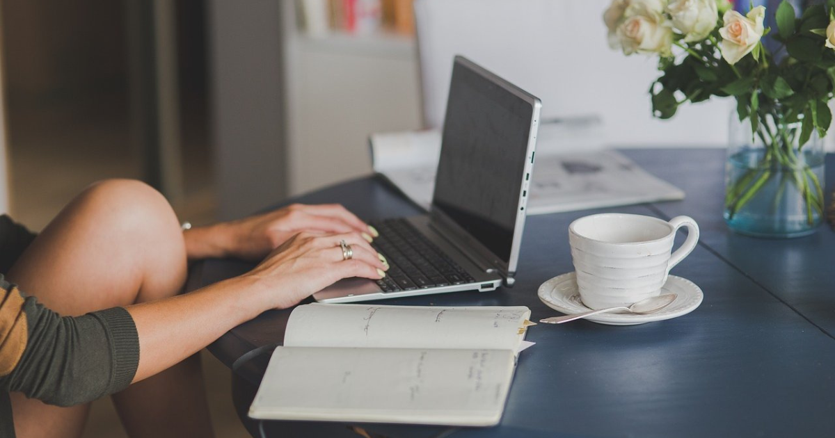 Womans hands typing on a laptop with an open notebook and a coffee cup nearby.