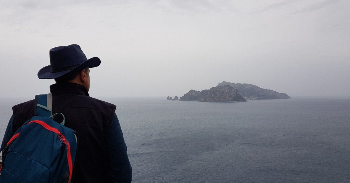View from behind a man with a backpack on the shore looking across water to rock island