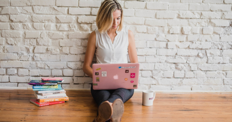 Woman sitting on the floor with a laptop computer next to her is a coffee mug and a stack of books