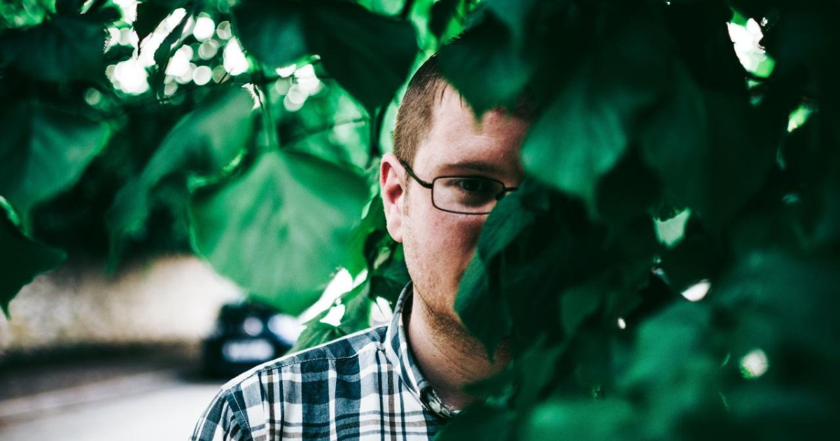 Man hiding behind the large leaves of a plant with only part of his face visible